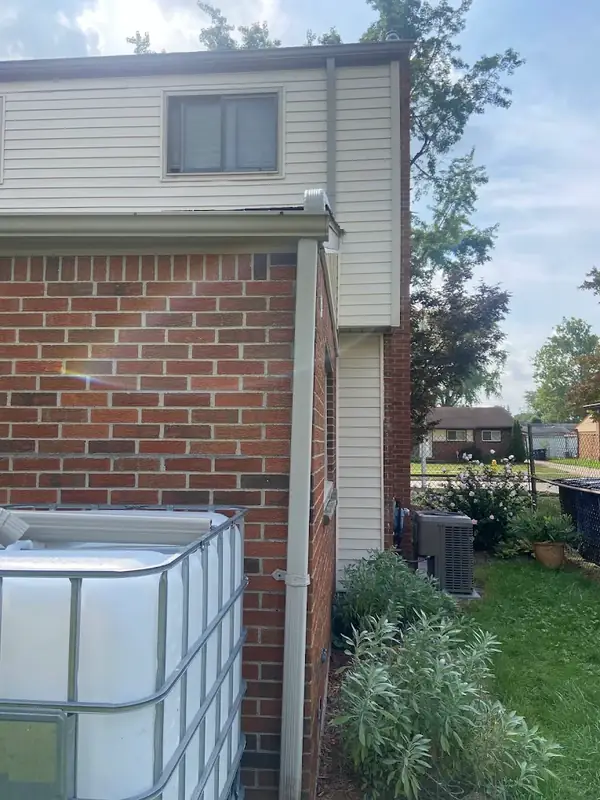 Tan downspout strapped to the brick corner of a two-story house, running from the gutter down past a white water tote