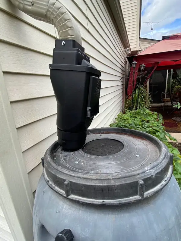 Close-up of a black downspout filter box connecting a tan downspout to the screened lid of a gray rain barrel
