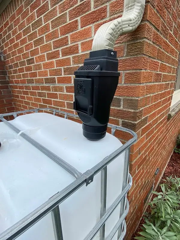 Black downspout filter mounted on top of a white caged water tote against a red brick wall