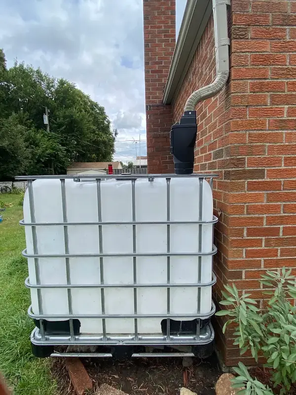 White caged water tote on a pallet beside a brick house, connected to a downspout that runs down from the gutter