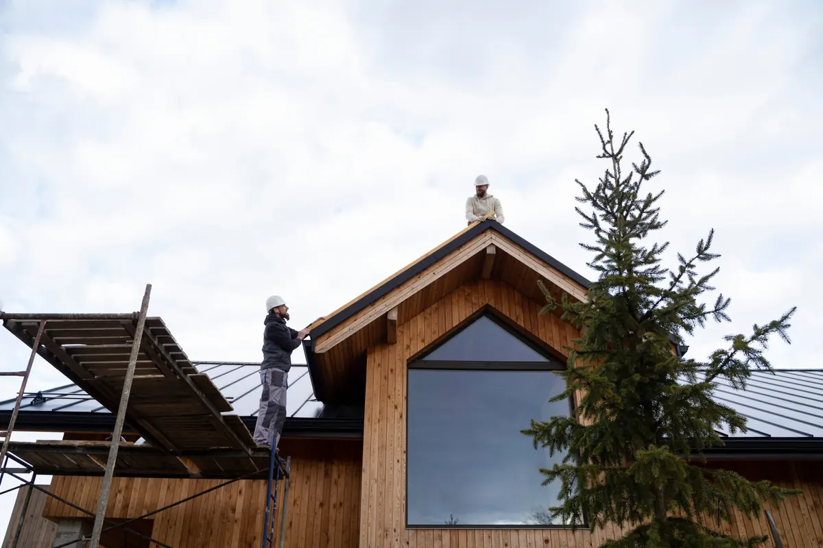 Two workers in hard hats at the roof edge of a wood-sided house, one on scaffolding at the fascia and one on the ridge
